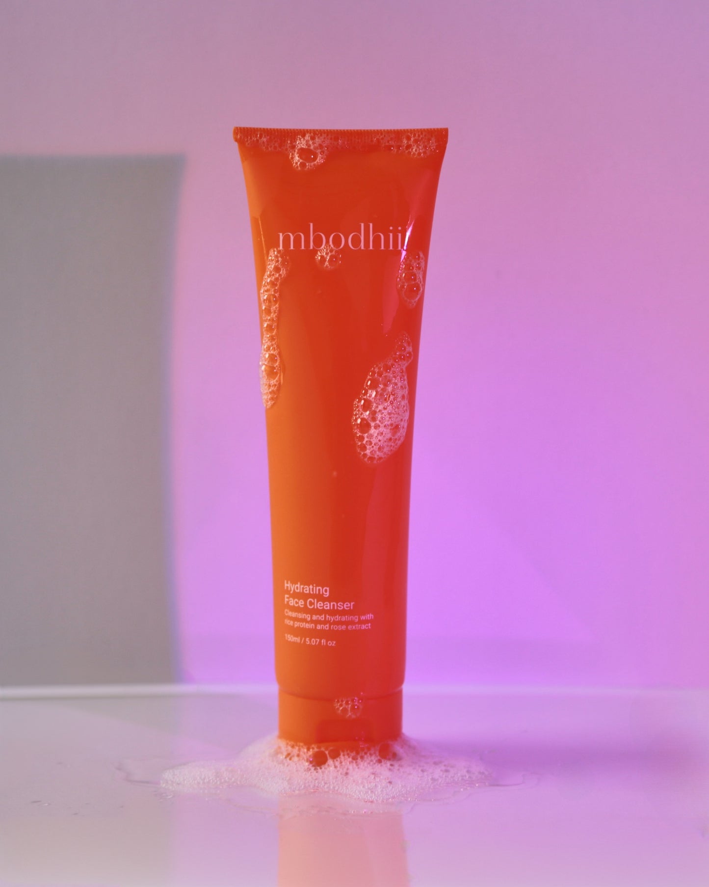Orange tube of mhoodhi facial cleanser on a white surface with a pink gradient background and some soapy suds